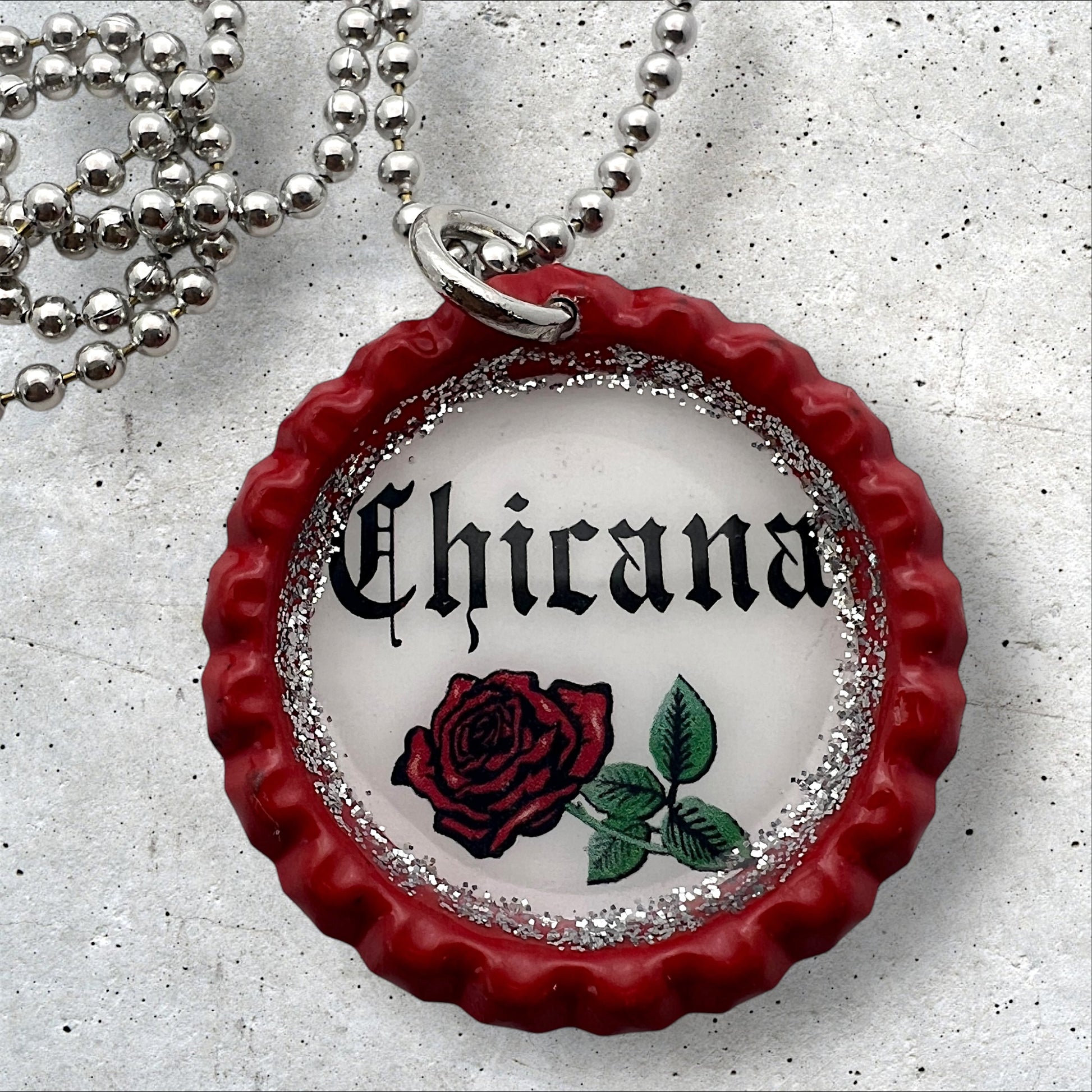 Photo of an Upcycled flattened red bottle cap pendant with the words chicana in the middle with a red rose underneath on a silver ball chain necklace. 