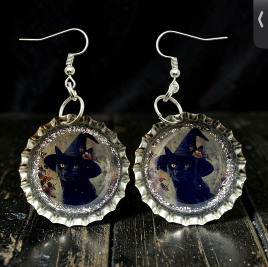 A pair of dangling earrings shaped like silver bottle caps, each featuring a black cat wearing a witch hat inside a glittery circular frame, shown on a dark wooden surface against a black background.