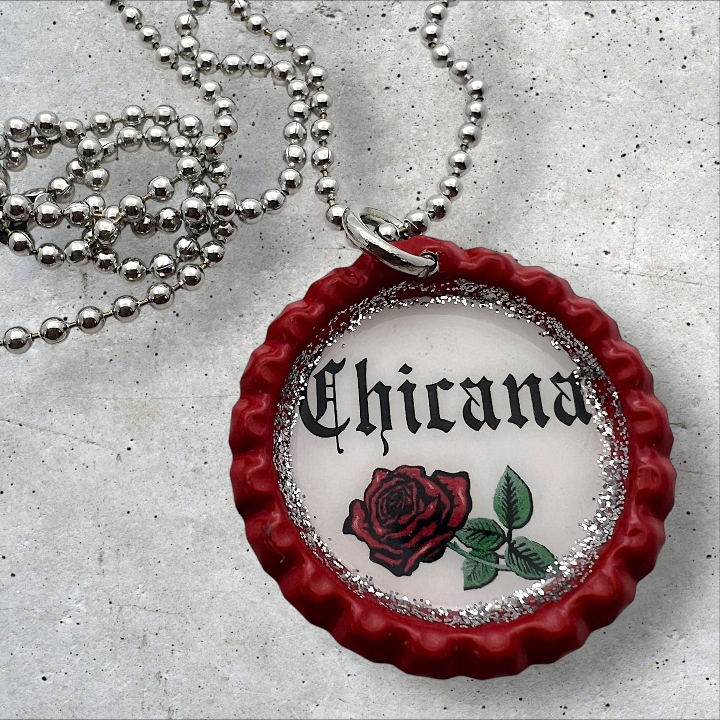 Photo of an Upcycled flattened red bottle cap pendant with the words chicana in the middle with a red rose underneath on a silver ball chain necklace. 