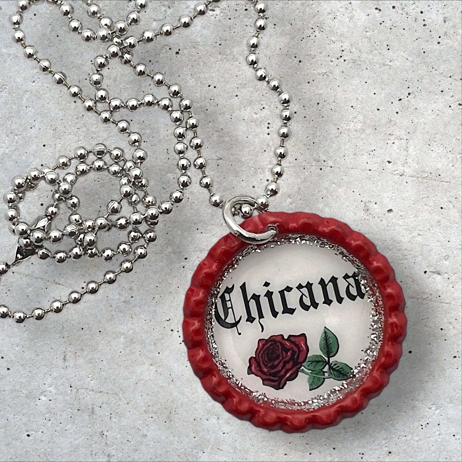 Photo of an Upcycled flattened red bottle cap pendant with the words chicana in the middle with a red rose underneath on a silver ball chain necklace. 